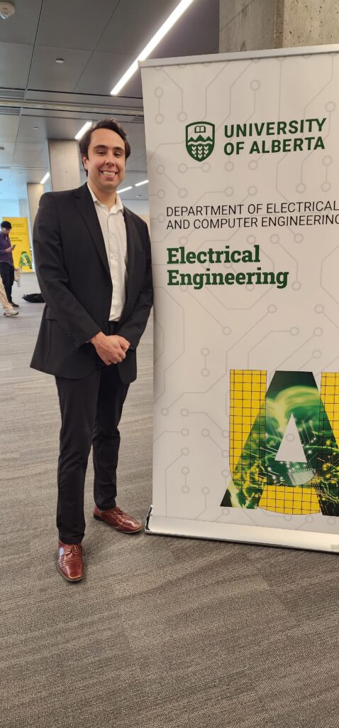Sean Piatt standing and smiling beside a large ‘Electrical Engineering’ department sign on campus.