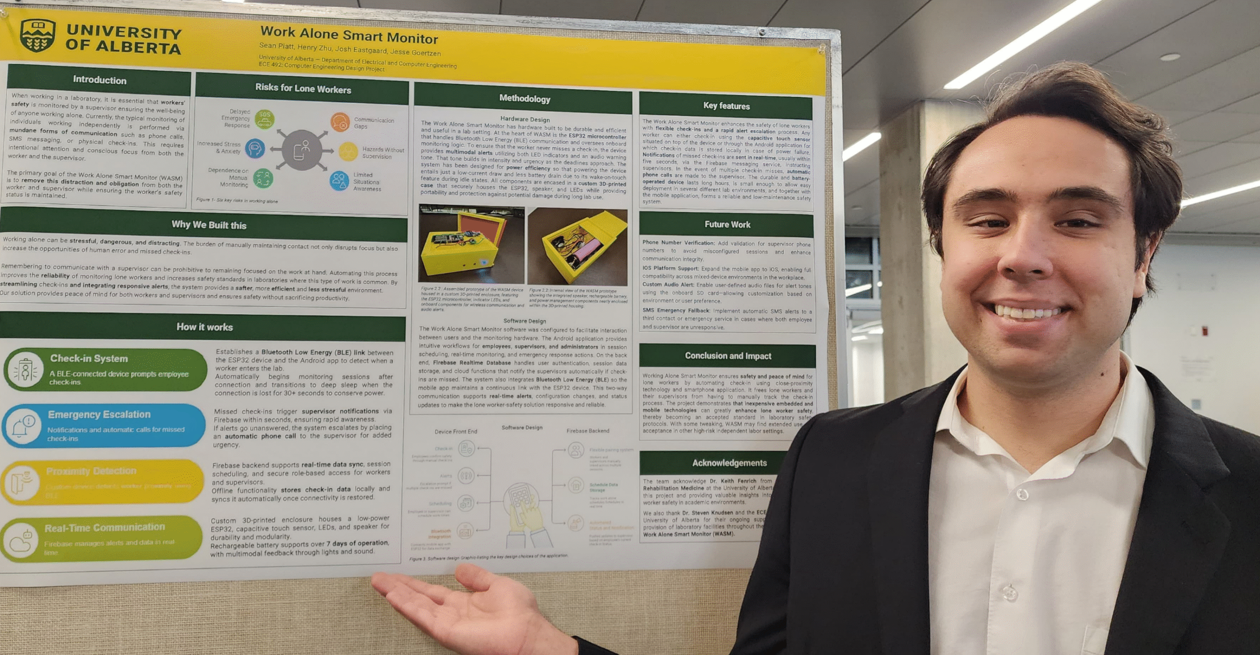 Sean Piatt pointing to his capstone project poster titled ‘Work Alone Smart Monitor (WASM)’ displayed on a conference board.